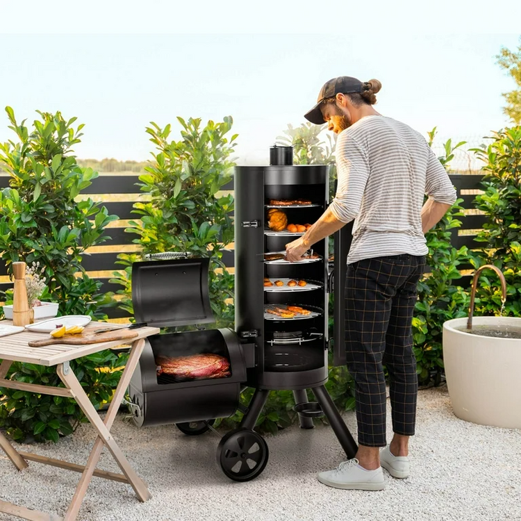 A man stands beside a large black outdoor smoker grill on a patio, placing trays of food into the upper chamber while another open compartment below cooks meat. The scene is set in a garden patio environment with greenery, a folding table, and a planter.